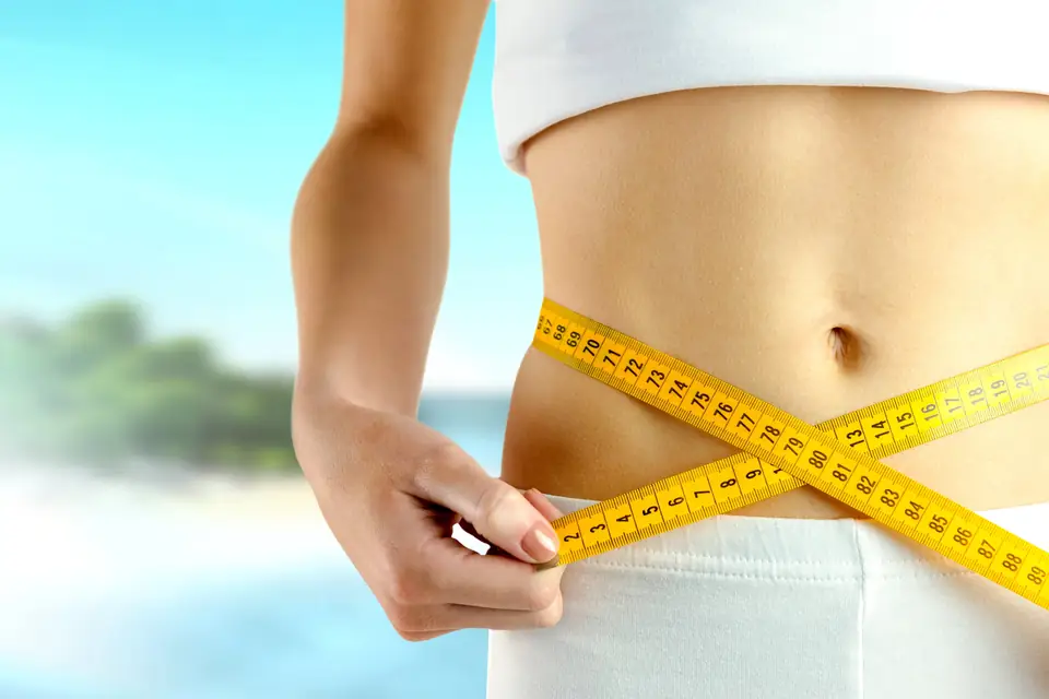 Beach background with a thin woman wrapping tape measure around her waist to illustrate weight loss.